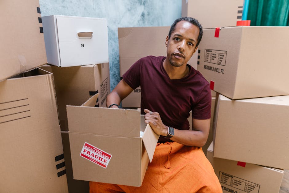 A man with dark skin and short curly hair, wearing a maroon t-shirt and orange pants, is sitting on the floor inside a room surrounded by stacked cardboard boxes used for house removals. He is holding an open box with one hand, while other boxes labeled 'fragile' and containing packing materials are visible around him. The room has a partially blue and beige wall in the background. The scene depicts the packing process during home relocation, with the man preparing items for transport. This image naturally relates to moving logistics and packing services offered by South Lambeth Removals, emphasizing careful handling of possessions during furniture transport and loading in preparation for residential moves.