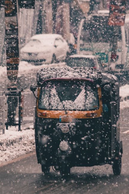 A three-wheeled tuk-tuk parked on a snow-covered street during winter weather, with snow visibly accumulating on its roof and windshield. In the background, blurred cars and trees are coated with snow, indicating recent snowfall. The vehicle appears stationary, and the street is lined with trees and buildings, with snow falling actively. This outdoor scene illustrates winter conditions affecting home relocation and furniture transport logistics, as seen in the context of booking removals slots in Lambeth during peak season through South Lambeth Removals.