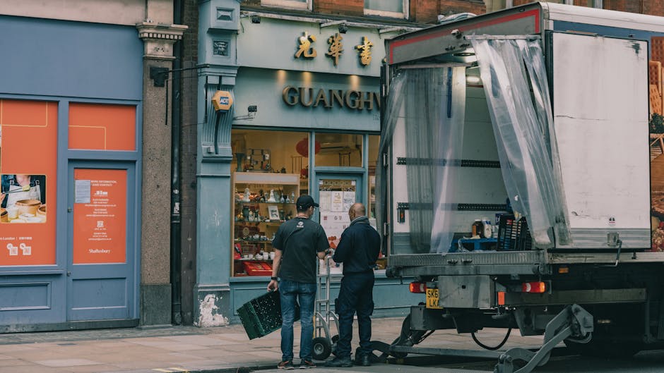 Two men are standing on the pavement outside a storefront with Chinese characters and the name 'GUANGH' displayed above the entrance, engaged in a conversation during the daytime. One man is holding a black crate, while the other is operating a hand truck or dolly. A partially loaded moving truck with an open rear door is parked nearby, with clear plastic wrapping draped over the accessible loading area. The truck is positioned close to the storefront, indicating an active loading process as part of a home relocation or furniture transport. The storefront's window displays various items, and the exterior features blue and grey tones with decorative architectural elements. The scene is illuminated by natural light, emphasizing the preparations involved in packing, moving, or furniture transport as managed by South Lambeth Removals, in the context of booking removals slots in Lambeth during peak season.