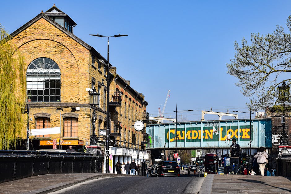 View of a busy street scene in Brixton Market during daytime, featuring a large brick building with arched windows and an iron fire escape, adjacent to a blue railway bridge with a prominent yellow and green 'Calden Lock' sign. The street is bustling with pedestrians, some crossing, others standing or walking along the pavement, and a variety of parked cars along the curb. In the foreground, a section of the pavement shows packing materials like cardboard boxes, plastic wrapping, and moving equipment such as trolleys and straps, indicating a home relocation or furniture transport activity. The scene is illuminated by natural daylight, with trees partially visible on the left and right edges, and construction cranes can be seen in the background, suggesting ongoing development nearby. South Lambeth Removals specializes in house removals and loading zone services, and is subtly reflected in the context of packing and moving logistics within this urban environment.