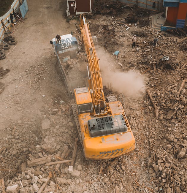 An aerial view of a large yellow construction excavator actively demolishing a concrete or brick structure at a building site, with dust and debris rising from the breaking process. The excavator is positioned on a rough, uneven ground surface covered with rubble, broken bricks, and debris. Nearby, a few workers are visible, observing or guiding the operation from a safe distance. To the left, there are construction materials and equipment, including a trolley loaded with boxes or packing materials, likely used for moving belongings or materials during a house relocation. The site is enclosed by temporary fencing with adjacent buildings and structures indicating an urban environment. The lighting suggests daytime, with clear weather conditions. The scene captures the intensive process of building deconstruction, relevant to clients interested in professional removals, packing, and planning logistics for home or commercial move activities, as seen on South Lambeth Removals' page about Brixton Market flat removals and loading zone advice.