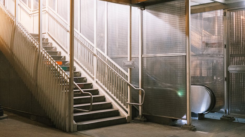 A staircase with metal handrails and steps covered in dark non-slip material, leading up from a dimly lit transit area inside a building. To the right of the stairs, there is an escalator housed within a metal and glass enclosure, with visible reflective surfaces and metallic panels. The environment appears to be part of an urban underground station or a similar public transport facility, with diffused lighting casting soft shadows. The space is clean and well-maintained, suitable for home relocation activities or furniture transport as part of the moving process, and suggests a setting where careful handling and navigation of stairs and escalators are necessary for moving household items efficiently. South Lambeth Removals often manages such logistics during residential moves in tight or challenging environments like this to ensure safe and professional home removal services.