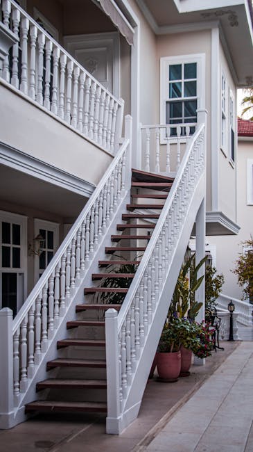 A white-painted outdoor staircase with wooden steps leading from the sidewalk to the first-floor entrance of a residential property in daylight. The staircase features decorative balustrades and handrails, with potted plants placed at the base on the pavement. The building has white exterior walls, and windows with divided panes are visible, along with a balcony area at the top of the stairs. The scene captures a typical home with exterior access, highlighting the staircase’s structure in a well-maintained environment. The image is relevant for house removals and packing and moving services, illustrating a location where furniture and boxes may need to be navigated through narrow or tight stair access during a relocation process, as handled by companies such as South Lambeth Removals.