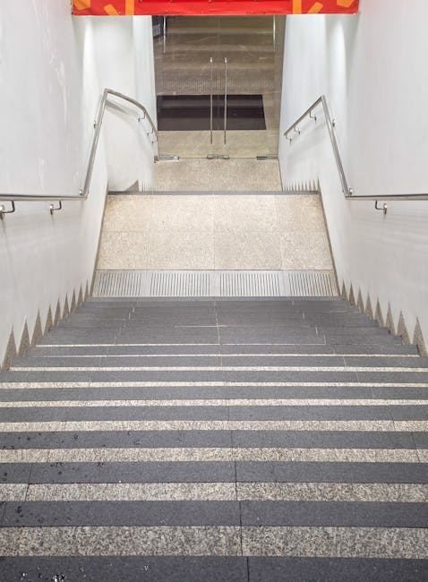 A staircase with metal handrails and steps covered in dark non-slip material, leading up from a dimly lit transit area inside a building. To the right of the stairs, there is an escalator housed within a metal and glass enclosure, with visible reflective surfaces and metallic panels. The environment appears to be part of an urban underground station or a similar public transport facility, with diffused lighting casting soft shadows. The space is clean and well-maintained, suitable for home relocation activities or furniture transport as part of the moving process, and suggests a setting where careful handling and navigation of stairs and escalators are necessary for moving household items efficiently. South Lambeth Removals often manages such logistics during residential moves in tight or challenging environments like this to ensure safe and professional home removal services.