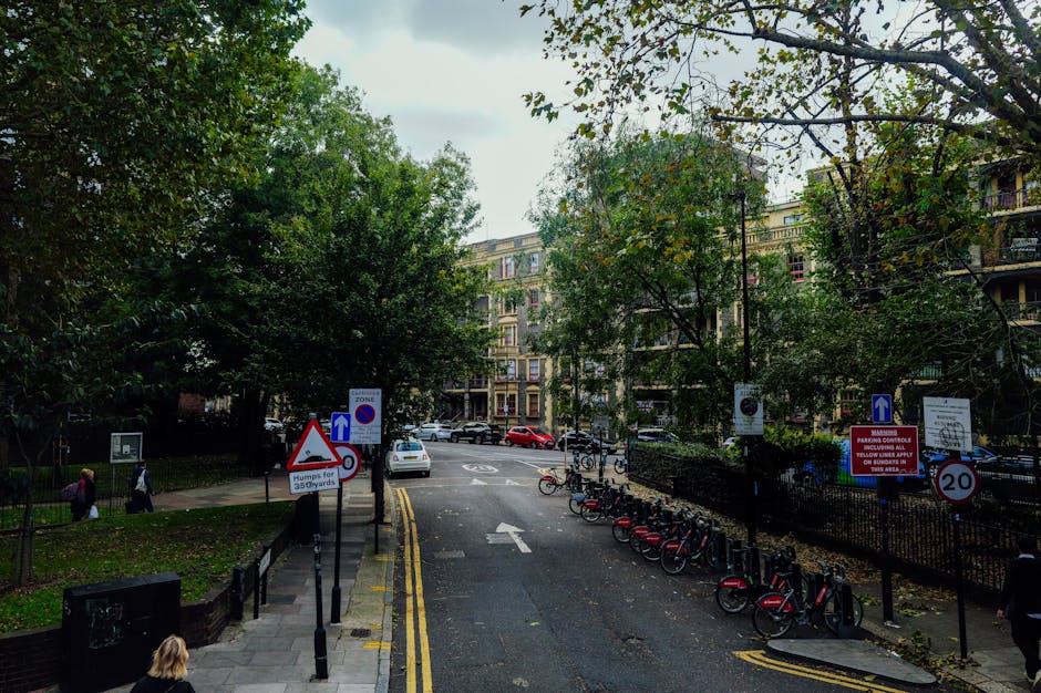 A street scene in Kennington with a view of residential buildings, trees, and parked bicycles. In the foreground, there are multiple traffic signs including a triangular caution sign, a parking zone indicator, and a 20 mph speed limit sign. The street is partially blocked by a black metal fence surrounding a green space with pedestrians walking on the pavement. Several bicycles are parked along the curb, and a white car is visible on the street. The scene is captured during daylight with natural lighting, and the trees have green leaves, indicating a season in early autumn. This image illustrates urban surroundings relevant to house removal planning, such as parking restrictions and loading areas, as seen in services provided by South Lambeth Removals for home relocation and furniture transport within a city environment.