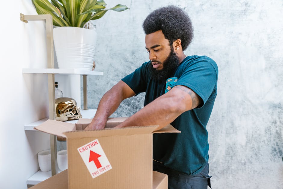 A man with dark curly hair, a beard, and wearing a navy blue t-shirt is seen packing items into a cardboard moving box. He is standing indoors in front of a light textured wall, with a white shelving unit behind him that holds a large potted plant with broad green leaves, a decorative gold skull, and some white cups. The man is placing his hands inside the open box, which is placed on a table or counter, and the box has a red 'CAUTION THIS SIDE UP' sticker on the front. The scene depicts the process of home relocation or packing and moving furniture and belongings, with sufficient lighting highlighting the packing materials and the man's focus on the task. South Lambeth Removals, a professional removals service provider, may assist with such packing and transportation tasks as part of house removals or furniture transport services.