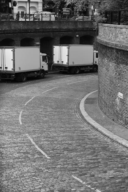 A black and white photograph depicting a curved cobblestone driveway with three closed moving trucks parked side by side, partially under an arched brick bridge structure. The trucks are positioned on the paved area adjacent to an exterior brick wall, with some visible packing materials and furniture, including cardboard boxes, wooden crates, and fabric-covered items, placed on the ground near the vehicles. The scene appears to be part of a home relocation or furniture transport operation, with the trucks ready for loading or unloading. The background shows a residential area with balconies and trees, indicating a city or town environment, and the lighting suggests daylight conditions. The image visualizes the logistical aspects of house removals, including vehicle positioning and packing in preparation for a move, aligned with the services offered by South Lambeth Removals.