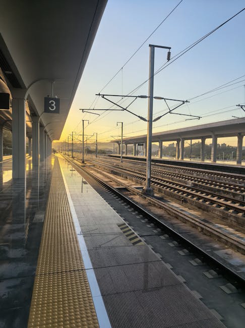 View of Vauxhall Station platform during early morning with a clear sky and rising sunlight illuminating the scene. The platform features a yellow tactile paving strip along the edge, alongside metal safety barriers and support columns. Overhead, electrical wires run parallel to the train tracks, supported by tall metal poles. The station's sheltered area extends to the left, with empty space indicating a quiet, pre-rush hour period. Nearby train tracks lead into the distance, with a series of overhead bridges and elevated roads visible beyond the platforms. The scene captures a calm moment suitable for a home relocation or furniture transport process, with emphasis on the station environment prior to an early morning move, as provided by South Lambeth Removals.