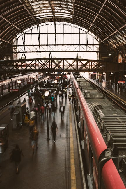 Inside Vauxhall Station during daytime with a high arched glass and metal ceiling allowing natural light to fill the spacious interior. The station platform is filled with numerous blurred figures of travelers, some walking, others waiting or moving toward the trains, which are visible alongside the platforms. A red and white train is parked on the right side, with sliding doors open for boarding. The floor is made of dark tiles, and there are yellow safety lines along the edge of the platform. Overhead, a bridge with a metal railing crosses the station, supporting various signage and lighting. The scene captures a busy moment typical of train stations during peak hours, providing context relevant to household or corporate home relocation processes, including logistics and transport considerations, which are supported by South Lambeth Removals' expertise in removals and moving services.